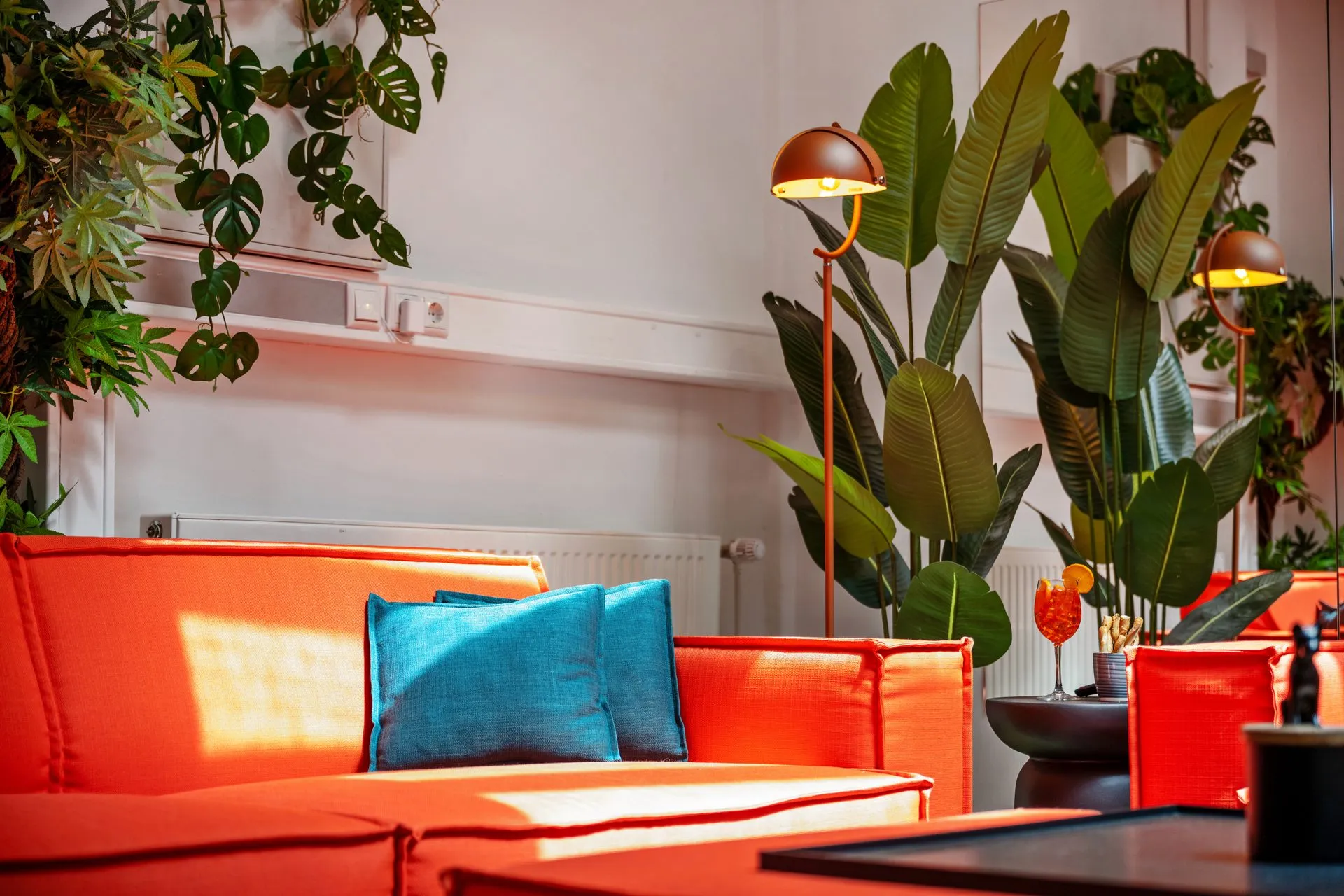Orangefarbenes Sofa mit türkisem Kissen zwischen Zimmerpflanzen und Stehleuchte